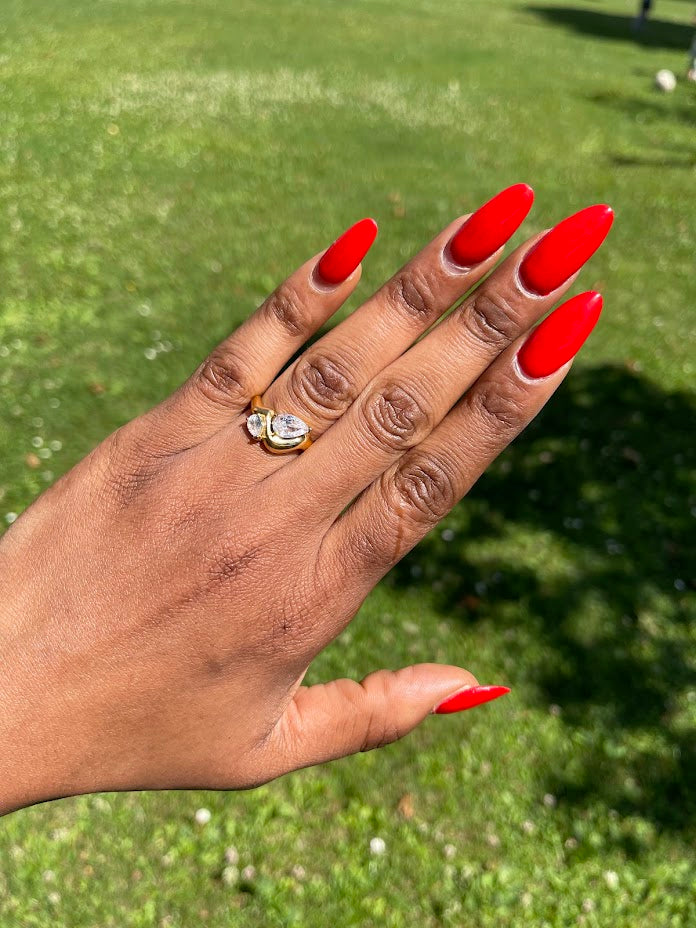 Hand wearing Selma Twin Pear-Cut Stone Ring with two pear-cut stones and wavy 18k gold-plated band against a green outdoor background.